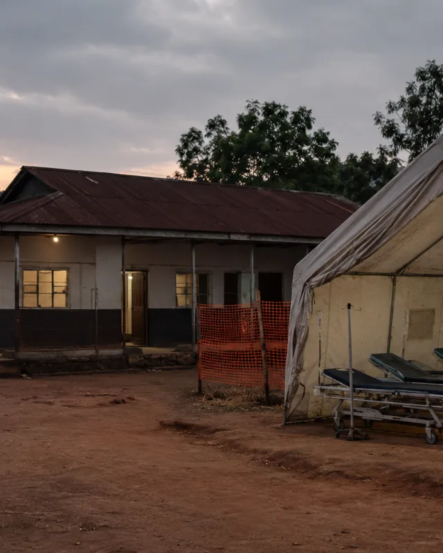 Exterior de una clínica de salud rural en la República Democrática del Congo al atardecer, camillas vacías bajo una carpa, tono documental