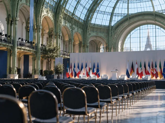 Sala de exposiciones parisina de cristal y hierro preparada para una cumbre internacional, con filas de sillas vacías frente a un escenario y banderas desenfocadas al fondo bajo la luz del techo acristalado