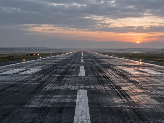 Pista de aeropuerto vacía vista desde el nivel del suelo al amanecer, con asfalto mojado, marcas de neumático y luces de borde perdiéndose en la bruma bajo un cielo encapotado